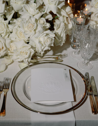 A formal dining table setting with white roses, crystal glassware, a place card, and silver cutlery on a white tablecloth.