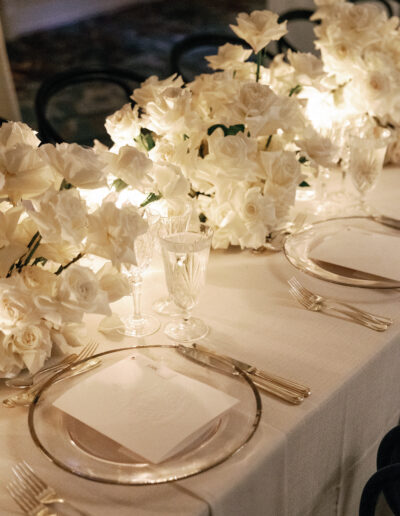 Elegant table setting with white roses, clear glassware, and gold-rimmed plates on a white tablecloth, softly lit.