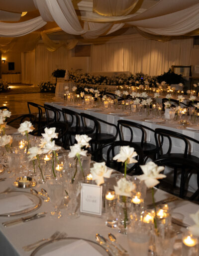 Elegant banquet hall with long tables set for an event. White flowers, candles, and black chairs adorn the setting, under draped fabric ceiling.