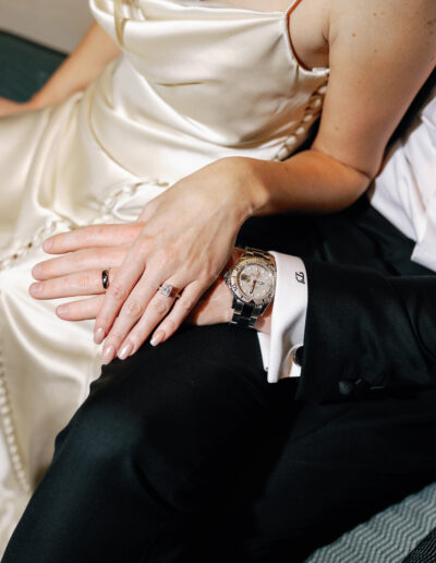 A couple in formal attire sit closely on a couch. They gently hold hands, showcasing their rings. The focus is on their hands and clothing details.