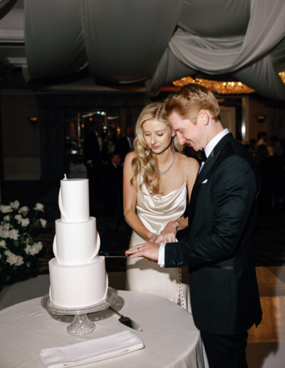 A couple in formal attire cuts a three-tiered white wedding cake together under a draped ceiling.