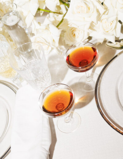 Two cocktail glasses filled with a brown liquid are set on a table with white roses and a clear crystal glass nearby.