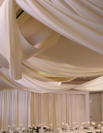 A decorated event space with draped white fabric on the ceiling, round tables set with white flowers, glassware, and black chairs.