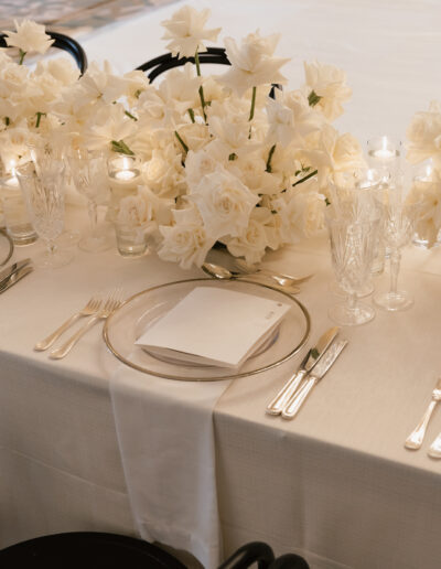 Elegant table setting with white roses, crystal glassware, silver cutlery, and white napkins on a table draped in a white cloth.