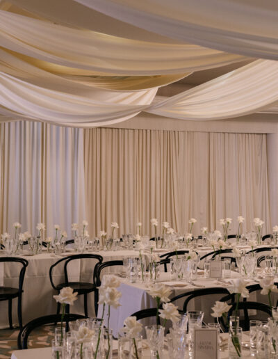 Elegant banquet hall with white draped ceiling, set tables with white flowers, glassware, and black chairs.