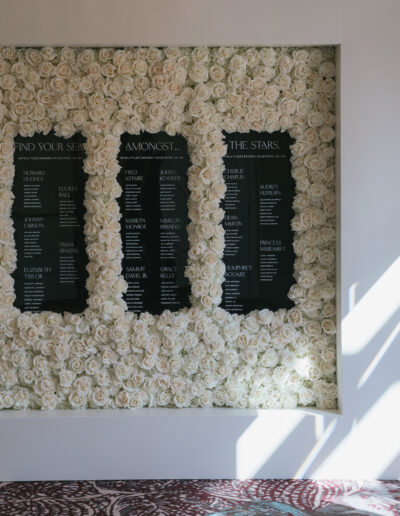 A wall covered in white roses with three black panels listing names in white text. Sunlight casts shadows on the right side of the wall.