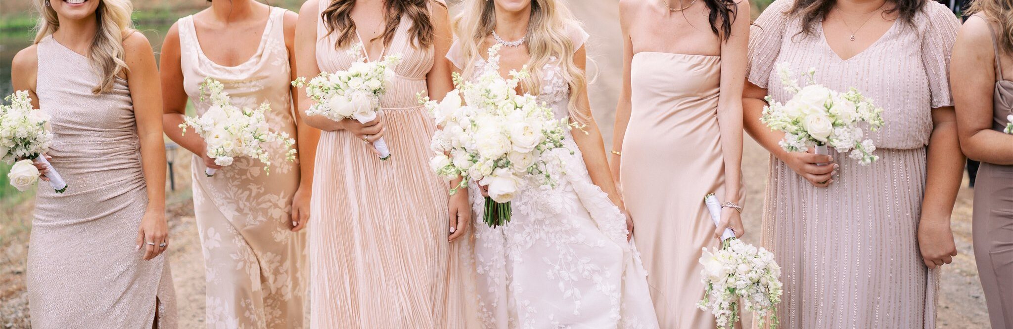 Six women in pastel dresses stand outdoors, holding white bouquets. They are smiling with a landscape of trees and a vineyard in the background.