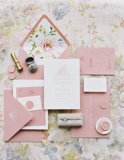 A floral-themed wedding invitation set with pink envelopes, invitation, cards, wax seals, a ring, stamp, and pen displayed on a floral patterned cloth background.