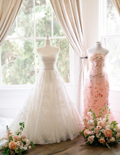 Two mannequins display elegant gowns, one white and one pink, surrounded by floral arrangements in front of large windows with light curtains.