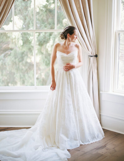 Bride in a white wedding gown stands by a large window with light curtains, looking to the side.