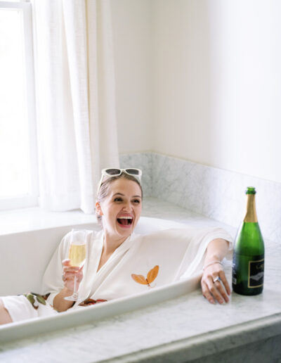 A woman in a bathrobe relaxes in a marble bathtub, holding a glass of champagne, with a bottle nearby, smiling broadly.