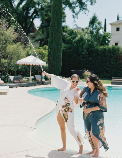 Two people in robes stand by a pool, one holding a champagne bottle spraying liquid, while the other smiles and watches.