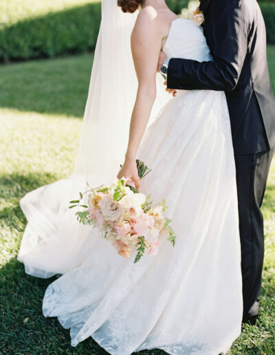 A bride and groom embrace outdoors. The bride holds a bouquet of flowers and wears a strapless white gown and veil. The groom is in a dark suit.