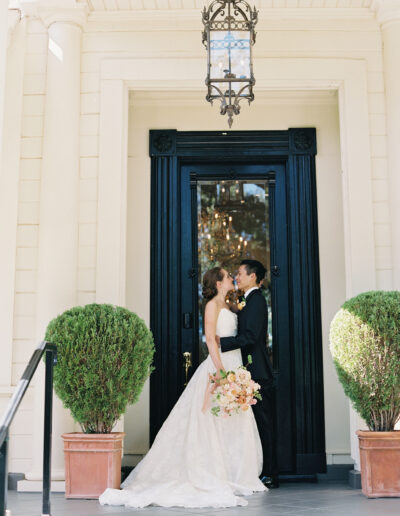 A bride and groom stand under a lantern at the entrance of a building, surrounded by potted plants. The bride holds a bouquet while they face each other.