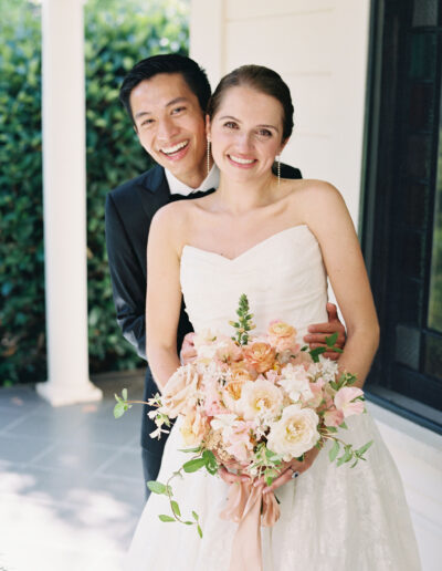 A couple in wedding attire smiles, with the bride holding a bouquet of pink and white flowers, standing on a porch.