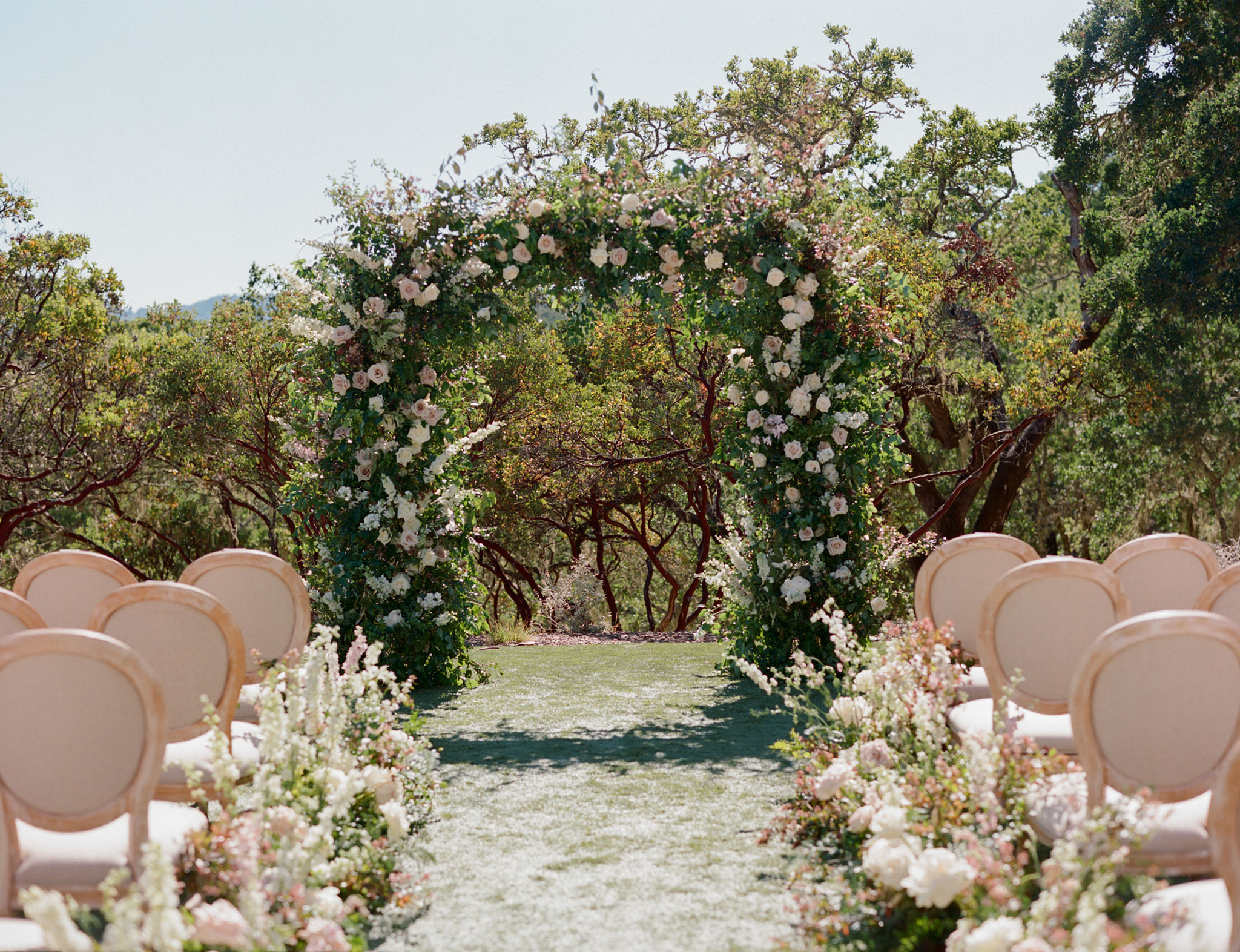 Outdoor wedding setup with a floral arch and rows of chairs on a grassy aisle lined with flowers. Trees and clear sky in the background.