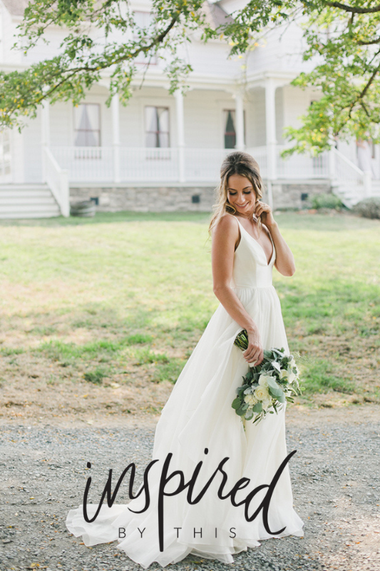 Bride in a white gown holding a bouquet outside a large white house on a sunny day.