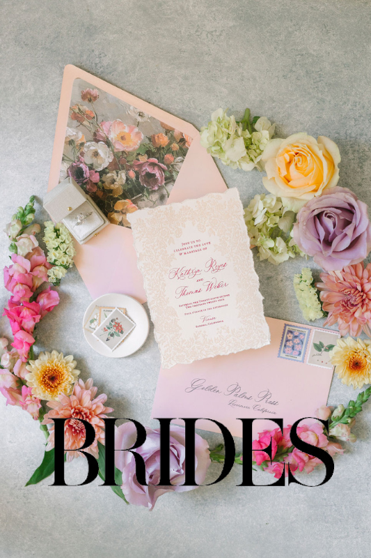 Wedding invitation set with floral designs, a ring box, and floral stamps surrounded by colorful flowers on a gray surface. "BRIDES" text at the bottom.