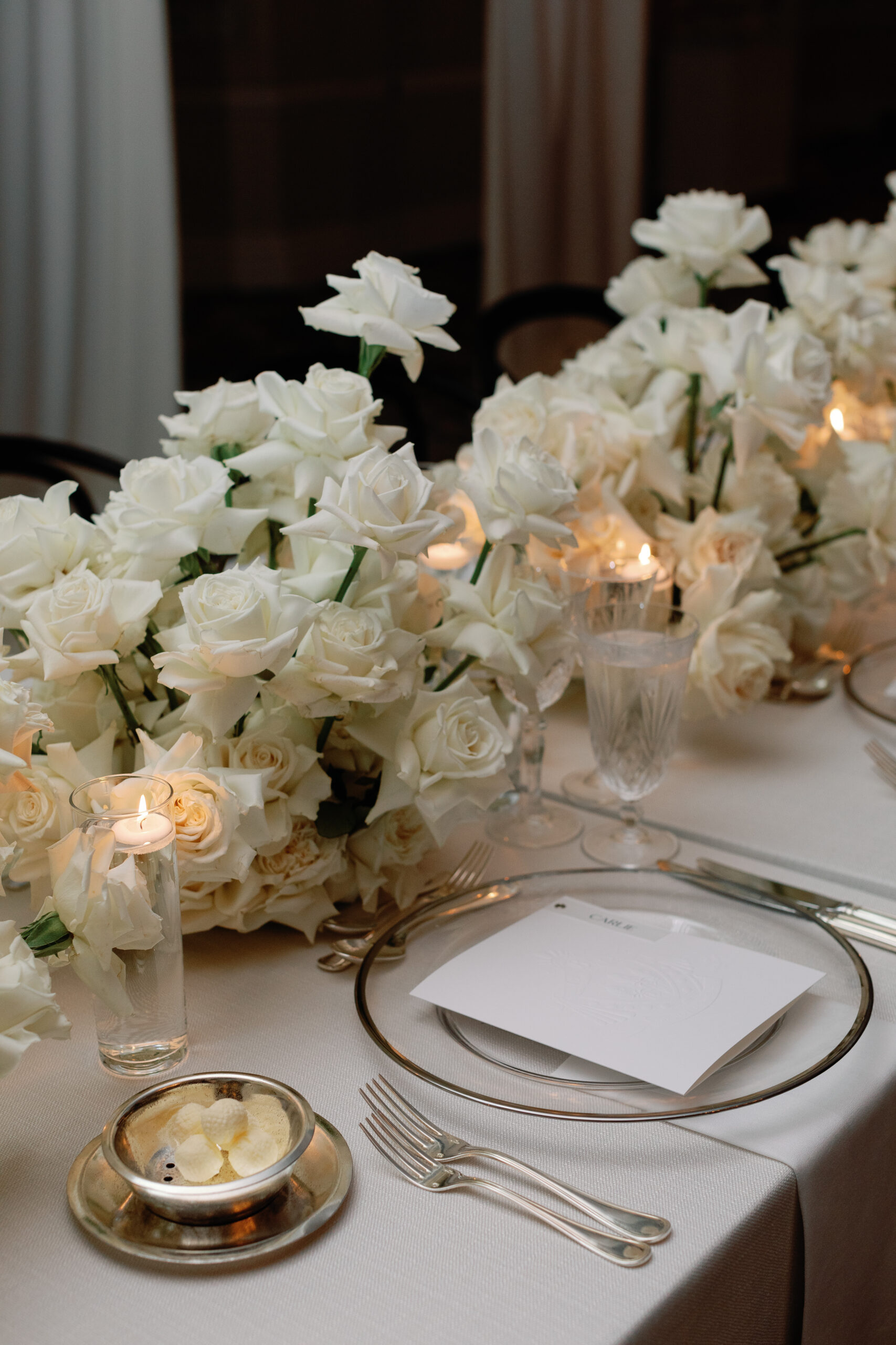 Elegant table setting with white roses, candles, and an empty plate with cutlery on a white tablecloth.