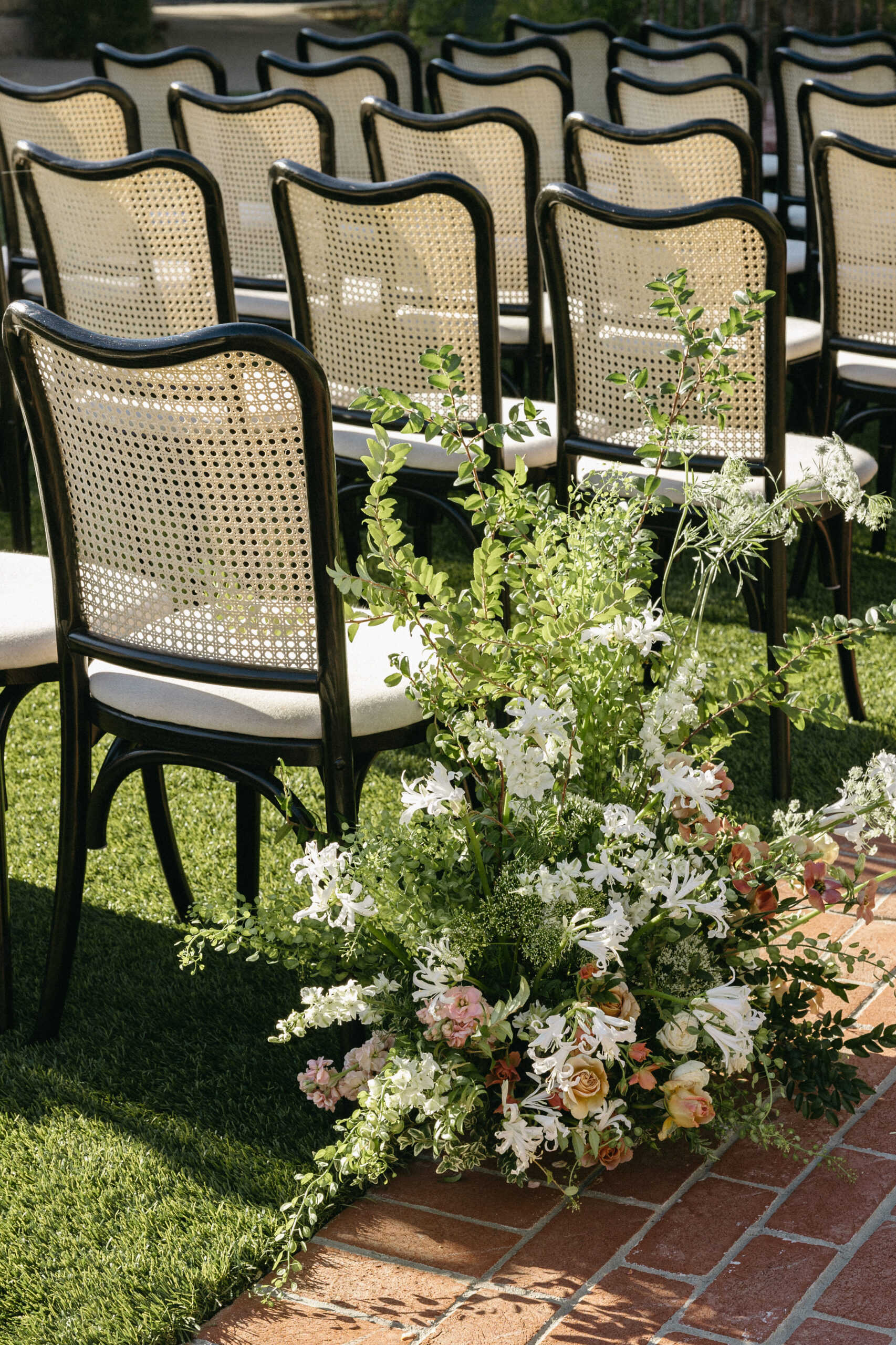 Rows of wicker-backed chairs on grass, with floral arrangements of greenery and white flowers along a brick pathway.