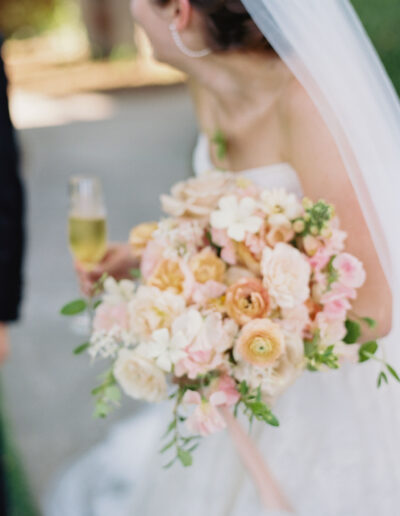 Bride holding a bouquet of pink, peach, and white flowers, and a glass of champagne.