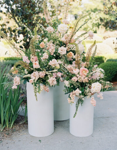 Three white cylindrical vases hold arrangements of pink and white flowers with greenery, set outdoors on a stone surface.