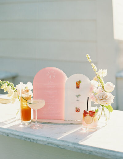 A menu with wedding drinks is displayed on a marble counter, featuring three cocktails in glasses with garnishes, surrounded by pink and white flowers.