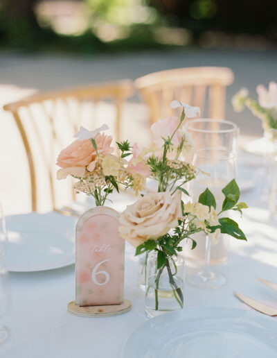 Table setting with a floral centerpiece featuring pale orange and cream roses, accompanied by a table number 6 sign, set on a white tablecloth with glassware and cutlery.