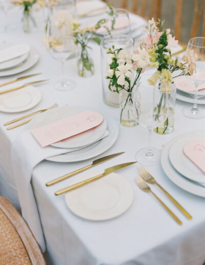 A table set for a formal meal with white plates, gold cutlery, pink menus, and small flower arrangements in glass vases.