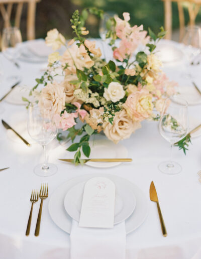 A round table with a white tablecloth is set with gold cutlery, a white plate, wine glasses, and a floral centerpiece of pastel flowers. A menu is placed on the plate.