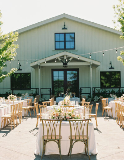 Outdoor wedding reception setup in front of a white barn. Tables are adorned with white tablecloths and floral centerpieces. String lights hang above, and wooden chairs surround the tables.