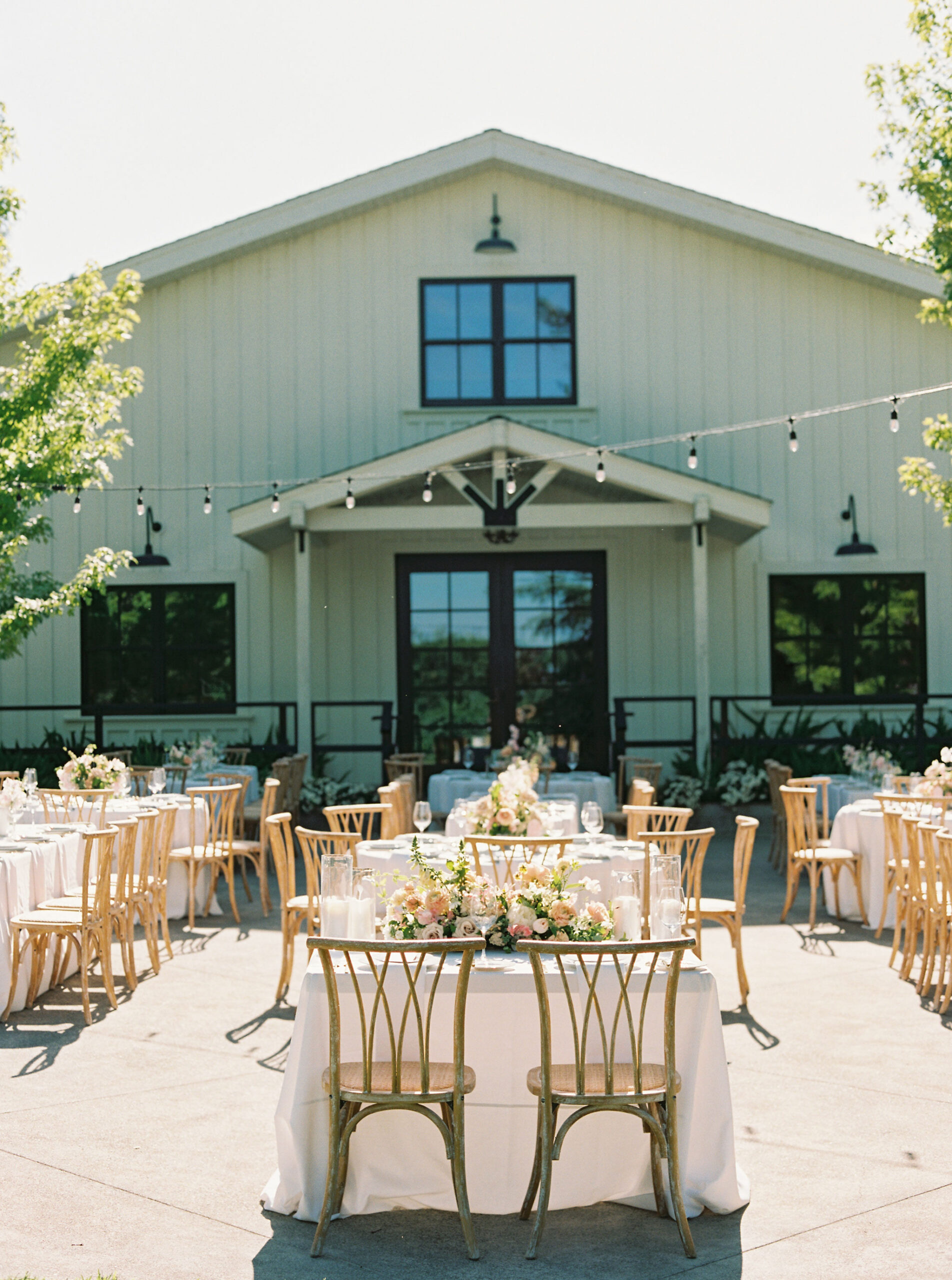 Outdoor wedding reception with wooden tables and chairs, decorated with floral centerpieces, in front of a rustic barn. String lights hang overhead.