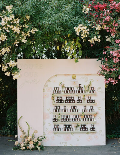 A decorative display with small bottles arranged on shelves against a floral backdrop, surrounded by blooming flowers and greenery. The words "You’re Our Jam" are written on the panel.