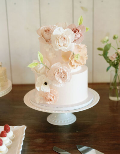 A two-tiered white cake with pink and white floral decorations on a cake stand, surrounded by desserts and flowers.