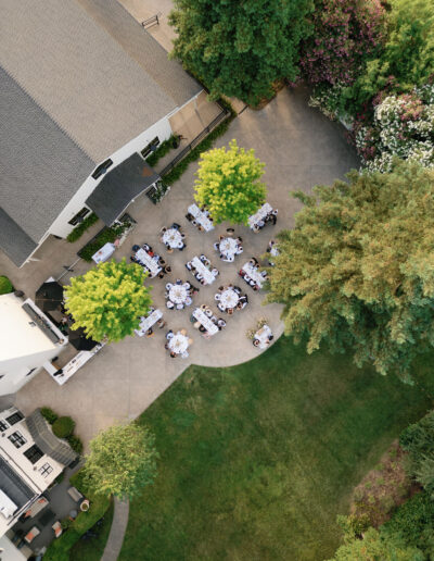 Aerial view of an outdoor wedding reception with several tables set for guests, situated on a concrete patio surrounded by green trees and landscaped lawn.