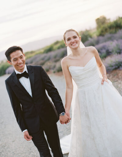 A bride in a white dress and a groom in a black suit hold hands while walking on a gravel path, with greenery in the background.