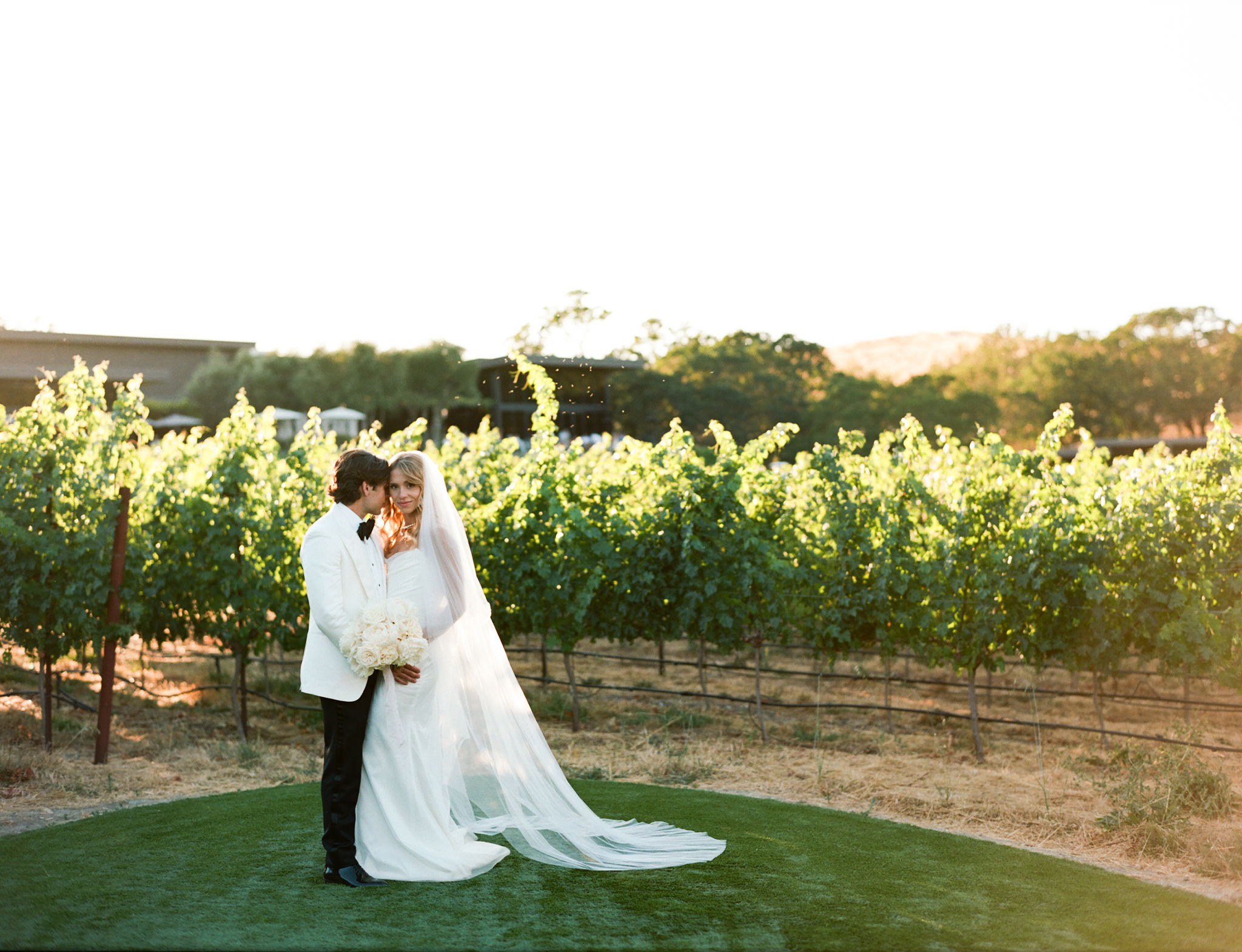 Bride and groom standing in a vineyard, the bride in a long white gown and veil holding a bouquet, while the groom wears a white jacket and black pants.