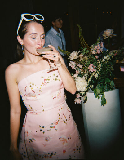 Woman in a floral dress sips a drink near a flower arrangement.