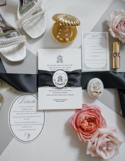 Wedding invitation set with a black ribbon, surrounded by white heels, pink flowers, pearl earrings, a gold ring, and a gold lipstick on a patterned surface.