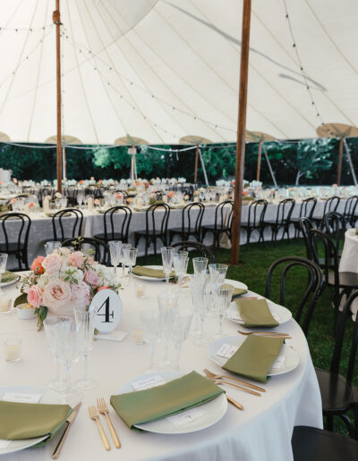 Outdoor event with white tent, round tables set with floral centerpieces, green napkins, glassware, and table numbers. Black chairs surround the tables on a grass lawn.