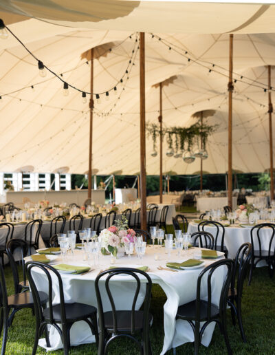 Outdoor event setup under a large tent with round tables, black chairs, white tablecloths, floral centerpieces, and string lights above.