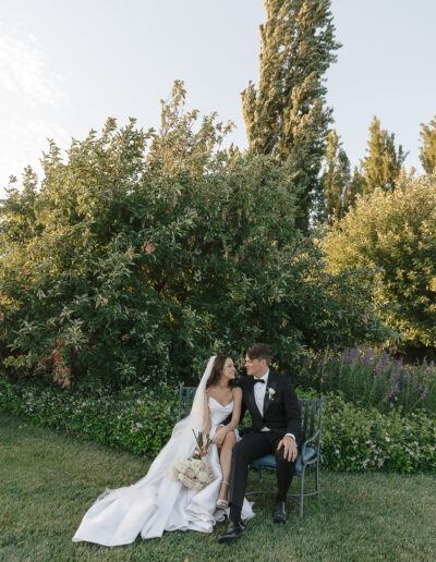 A bride and groom sit on a bench in a garden. The bride wears a white dress and holds a bouquet, while the groom wears a black suit. They are looking at each other.