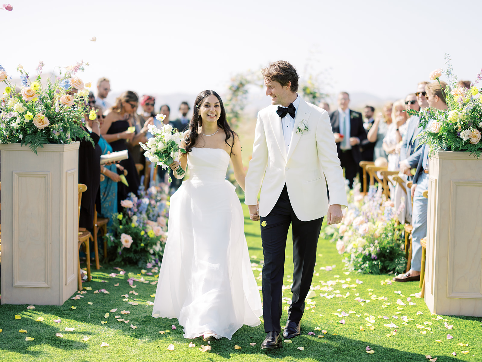 A couple, dressed in wedding attire, walks down an outdoor aisle lined with flowers, orchestrated flawlessly by a Napa wedding planner, surrounded by delighted guests.