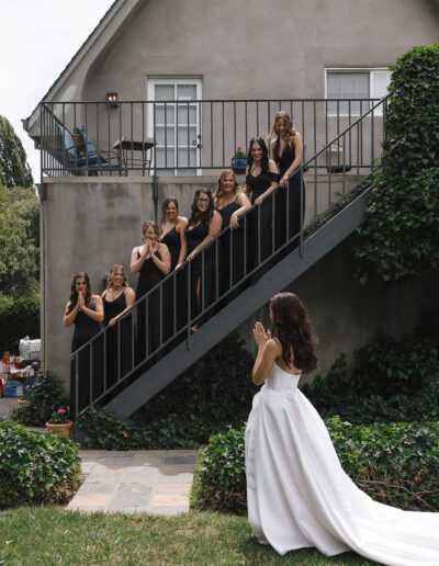 A bride in a white dress stands at the bottom of outdoor stairs, while seven bridesmaids in black dresses pose along the steps, looking towards her.