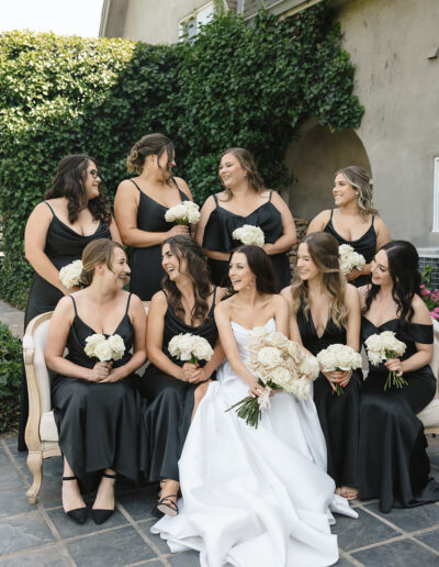 A bride in a white dress sits with her bridesmaids in black dresses, all holding white roses, in front of a vine-covered wall.