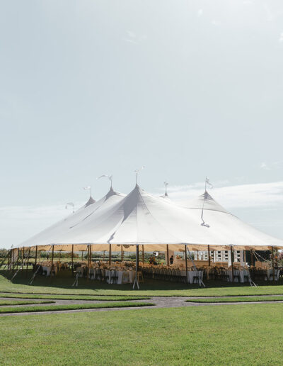 A large white tent is set up in the middle of a grass field, surrounded by trees and a clear sky.