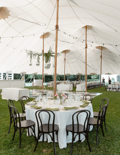 An outdoor event tent with a round table set for dinner, surrounded by dark wooden chairs on a grassy area. The tent is decorated with plants and string lights.