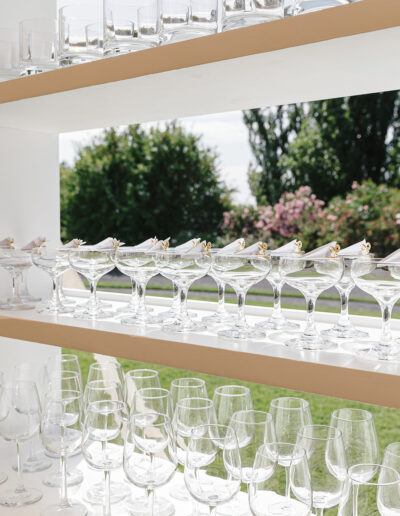 Rows of empty wine and cocktail glasses arranged on wooden shelves outdoors.