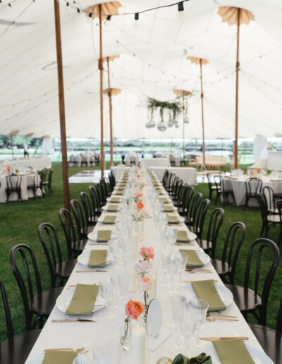 Long table set for an event under a white tent, featuring green napkins, glassware, and floral centerpieces with chairs on either side.