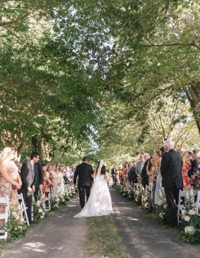 Bride and groom walking down an outdoor aisle lined with trees and guests on both sides.